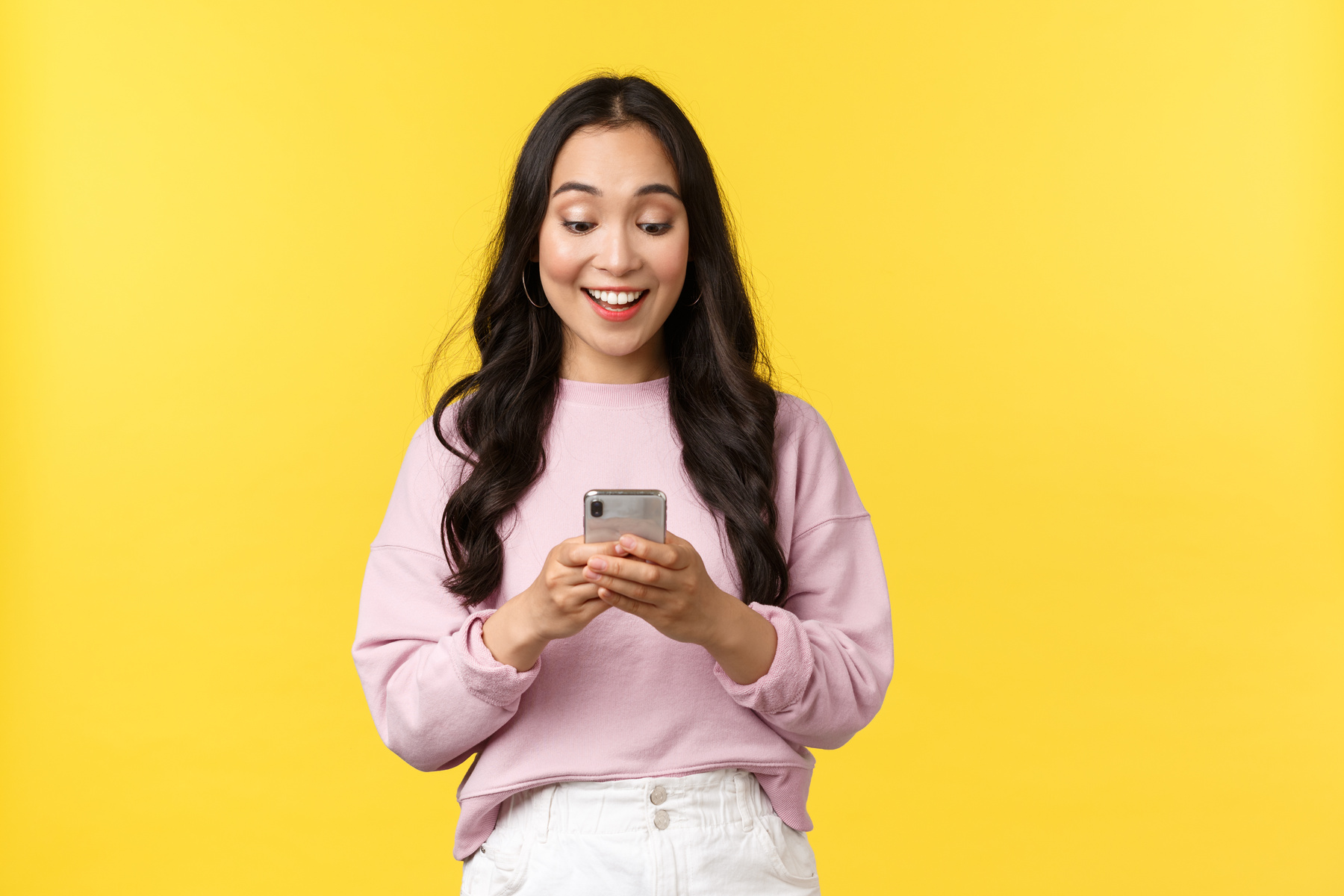 People Emotions, Lifestyle Leisure and Beauty Concept. Surprised and Happy Asian Girl Receive Great News over Phone, Looking at Mobile Display with Amazed Smile, Standing Yellow Background Rejoicing